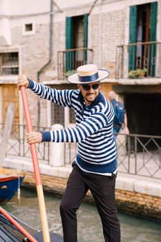 A smiling happy gondolier controls a gondola with tourists using a paddle. Classic blue-and-white striped jacket and hat is a historical image.