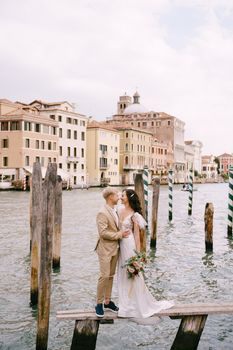 The bride and groom stand on a wooden dock for boats and gondolas, near striped green and white mooring poles, against the facades of the Grand Canal buildings.