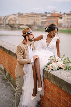 An African-American bride sits on a brick wall and a Caucasian groom hugs her. The embankment of the Arno River, overlooking city and bridges. Interracial wedding couple. Wedding in Florence, Italy.