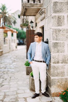 A man in a blue jacket with a bow tie stands in front of an ancient building, holding his hands in his pockets. High quality photo
