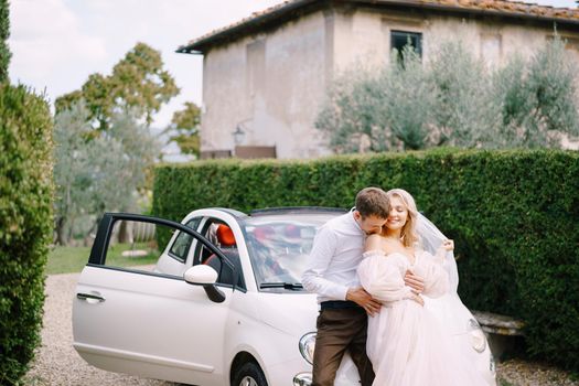 Groom kissing his bride on the dhoulder in front of a convertible at the old villa in Italy, in Tuscany, near Florence