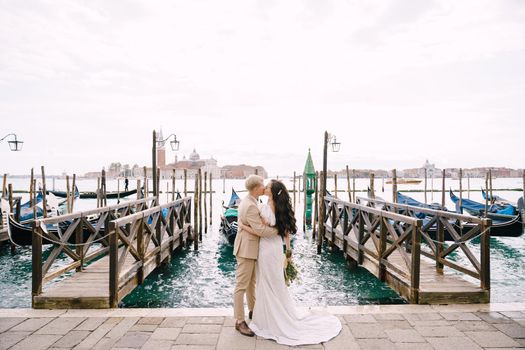 The bride and groom kiss on the gondola dock, cuddling, in Venice, near Piazza San Marco, overlooking San Giorgio Maggiore and the sunset sky. The largest pier for gondolas in Venice, Italy.