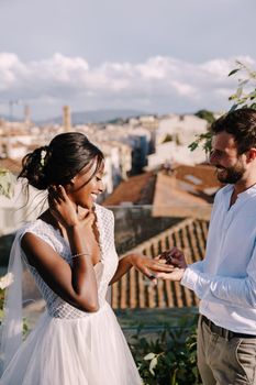 Interracial wedding couple - African-American bride and Caucasian groom. Groom puts a ring on brides finger. Destination fine-art wedding in Florence, Italy. Wedding ceremony on the roof of building