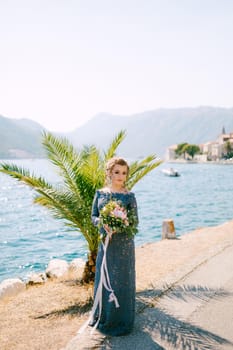 The bride in a stylish gray dress stands with wedding bouquets on the pier near old town of Perast. High quality photo