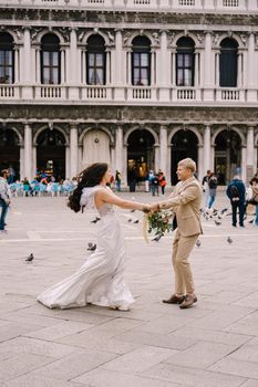 Wedding in Venice, Italy. The bride and groom are dancing among many pigeons in Piazza San Marco, against the backdrop of the National Archaeological Museum Venice, surrounded by a crowd of tourists.