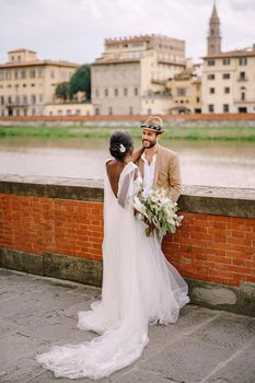 African-American bride and Caucasian groom stand hugging on the embankment of the Arno River, overlooking the city and bridges. Interracial wedding couple. Wedding in Florence, Italy.