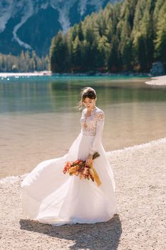 Beautiful bride in a white dress with sleeves and lace, with a yellow autumn bouquet of dried flowers and peony roses, at Lago di Braies in Italy. Destination wedding in Europe, at Braies lake.