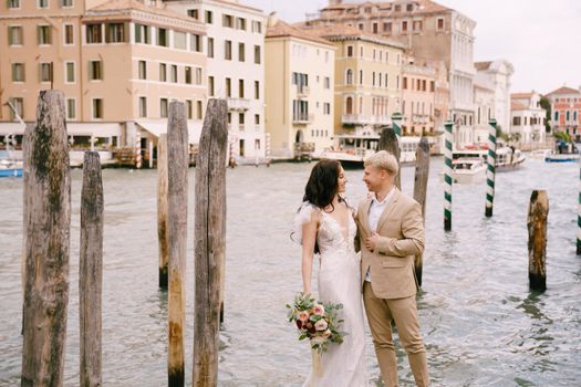 The bride and groom stand on a wooden dock for boats and gondolas, near striped green and white mooring poles, against the facades of the Grand Canal buildings.