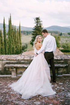 Wedding at an old winery villa in Tuscany, Italy. The wedding couple stands on the roof of an old winery, cuddling, standing with their backs in the frame.