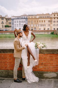 An African-American bride sits on a brick wall and a Caucasian groom hugs her. The embankment of the Arno River, overlooking city and bridges. Interracial wedding couple. Wedding in Florence, Italy.