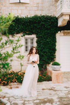 A bride with a bouquet in her hands stands at the wall of a house with a green liana and orange flowers in Perast . High quality photo