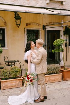 The bride and groom walk through the deserted streets of the city. Newlyweds kiss in the background of an old house.