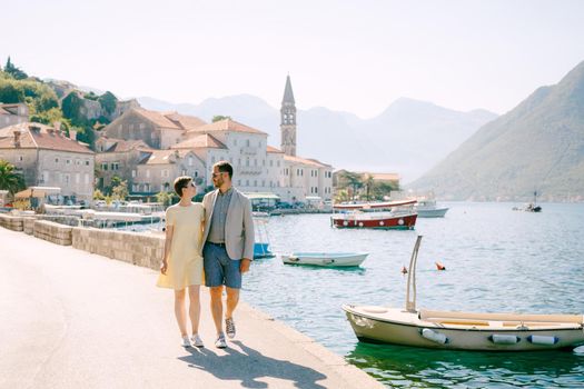 A man and a woman in sunglasses walk hugging each other on the pier near the boats in old town of Perast. High quality photo