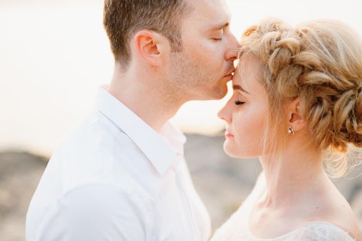 The groom gently kisses the bride on the forehead on the rocks by the sea, close-up. High quality photo