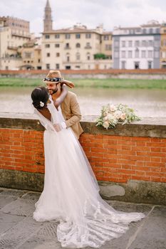 African-American bride and Caucasian groom stand hugging on the embankment of the Arno River, overlooking the city and bridges. Interracial wedding couple. Wedding in Florence, Italy.