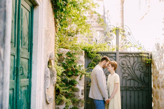 A man and a woman stand holding hands and a forged gate near an ancient building in Perast . High quality photo