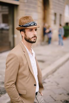 A young guy with a straw hat, a white unbuttoned shirt and a sand jacket, a small beard walks around of Florence.