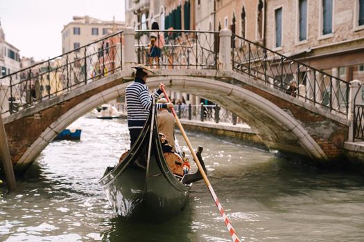 The gondolier rides the bride and groom in a classic wooden gondola along a narrow Venetian canal. The gondola swims under an arched bridge, a view from behind.