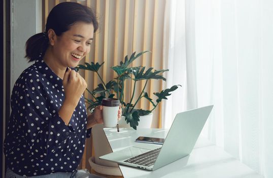 Business woman holding cup of tea and video calling via laptop talk to assistant in cafe. She is very happy to get the order on a video meeting. Work from home concept during Coronavirus pandemic.