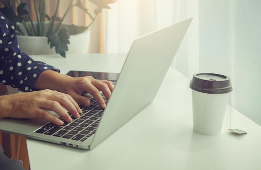 Young woman using laptop, communicates on internet with customer in home garden/greenhouse, reusable coffee/tea mug on table. Cozy office workplace, remote work, E-learning concept.