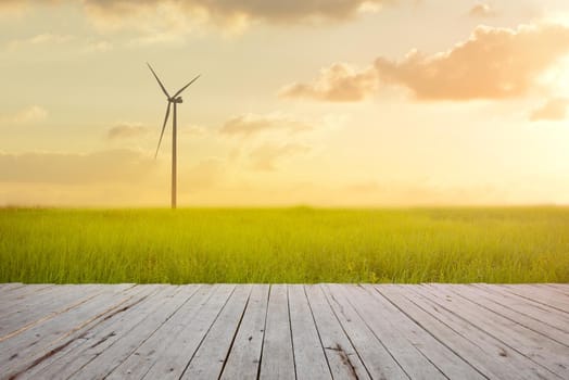 Wind turbine on green rice field against sunset background with plank wood foreground. Green energy concept.