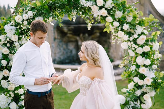 Wedding at an old winery villa in Tuscany, Italy. Round wedding arch decorated with white flowers and greenery in front of an ancient Italian architecture. The groom puts ring on the brides finger.