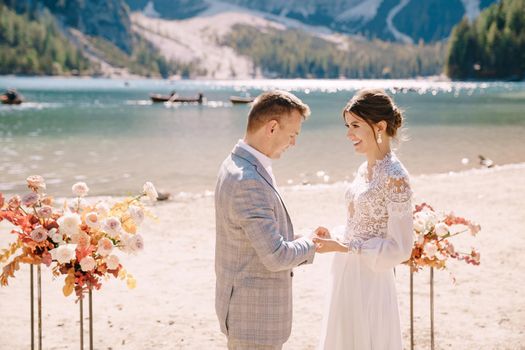The bride dons a ring to the groom at the venue for the ceremony, with an arch of autumn floral columns, against the backdrop of Lago di Braies in Italy. Destination wedding in Europe, at Braies lake.