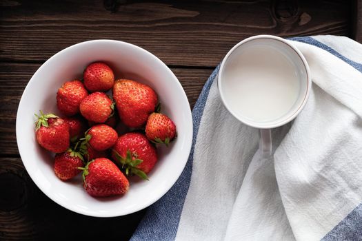 Natural ripe strawberries in a white bowl and milk in a mug on a dark wooden background. Top view.