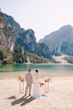 The bride and groom stand still for the ceremony, with an arch of autumn floral columns, against the backdrop of Lago di Braies in Italy. Destination wedding in Europe, at Braies lake.