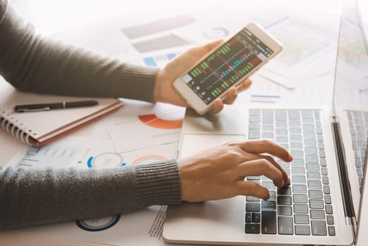 Businesswoman analyze business report graph and finance chart using smartphone and computer laptop at corporate office.