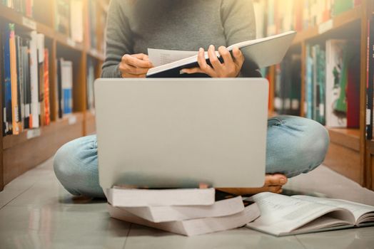 A woman sitting at library looking a book with laptop computer with blurred bookshelf background.