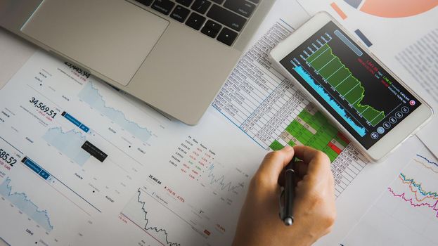 Businesswoman analyze business report graph and finance chart using smartphone and computer laptop at corporate office.