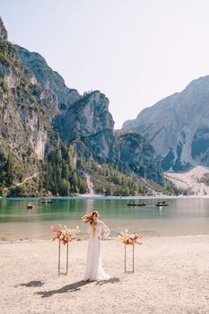 Beautiful bride in a white dress with sleeves and lace, with a yellow autumn bouquet against backdrop of arch for ceremony, at Lago di Braies in Italy. Destination wedding in Europe, at Braies lake.