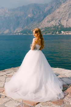 A beautiful blonde bride in a puffy white dress stands on a pier in the Bay of Kotor . High quality photo