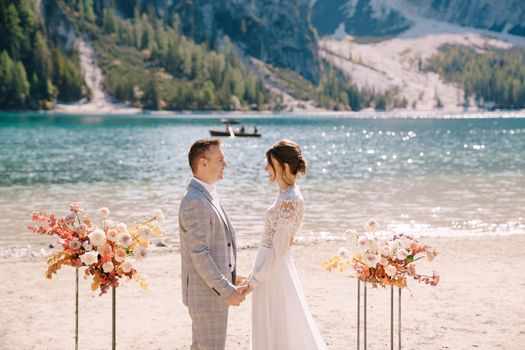 The groom dons a bride's ring, in place for the ceremony, with an arch of autumn floral columns, against the backdrop of Lago di Braies in Italy. Destination wedding in Europe, at Braies lake.