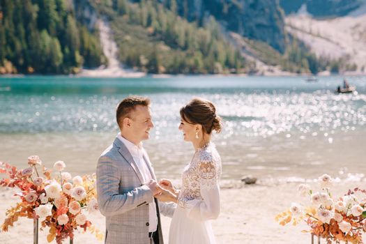 The bride and groom stand still for the ceremony, with an arch of autumn floral columns, against the backdrop of Lago di Braies in Italy. Destination wedding in Europe, at Braies lake.