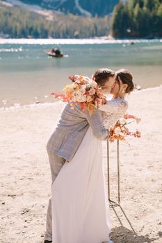 Newlyweds kiss on the spot for the ceremony, with an arch of autumn flower columns, against the backdrop of Lago di Braies in Italy. Destination wedding in Europe, at Braies lake.