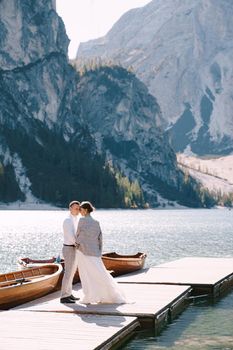 The bride and groom walk on a wooden boat dock at Lago di Braies in Italy. Wedding in Europe, at Braies lake. Newlyweds walk, kiss, cuddle against the backdrop of rocky mountains.