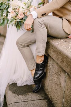 Close-up of the groom's legs in light short trousers and black leather shoes, the bride in a white dress with a bouquet in her hands.