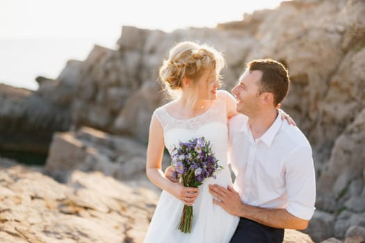 Bride with a bouquet of blue flowers and the groom hug at the rocks by the sea . High quality photo