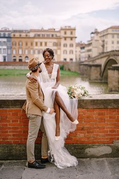 An African-American bride sits on a brick wall and a Caucasian groom hugs her. The embankment of the Arno River, overlooking city and bridges. Interracial wedding couple. Wedding in Florence, Italy.