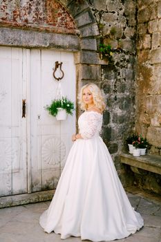 A bride stands in front of an old brick building near a white wooden door with flower pots in Perast . High quality photo