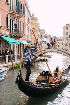 The gondolier rides the bride and groom in a classic wooden gondola along a narrow Venetian canal. The newlyweds are sitting in the boat, the captain pushes off the paddle.