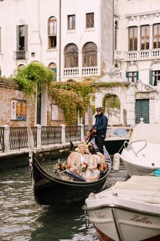 The gondolier rides the bride and groom in a classic wooden gondola along a narrow Venetian canal. The newlyweds sit in a boat and swim past other moored boats.
