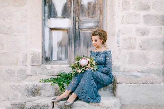 A bride in a stylish blue dress with a bouquet in her hands sits on the steps near an old wooden door . High quality photo