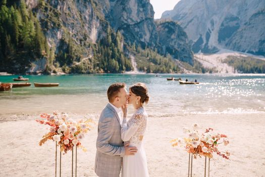 The groom dons a bride's ring, in place for the ceremony, with an arch of autumn floral columns, against the backdrop of Lago di Braies in Italy. Destination wedding in Europe, at Braies lake.