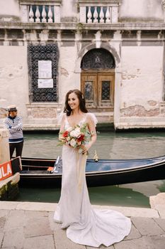A bride in a white dress, with a train, with a bouquet of white and red roses in her hands, stands on the pier near the moored gondola in a narrow Venetian canal.