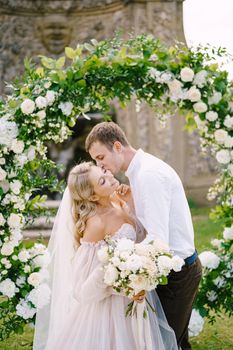 Wedding at an old winery villa in Tuscany, Italy. Round wedding arch decorated with white flowers and greenery in front of an ancient Italian architecture. The wedding couple is kissing.