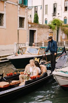 The gondolier rides the bride and groom in a classic wooden gondola along a narrow Venetian canal. The newlyweds sit in a boat and swim past other moored boats.