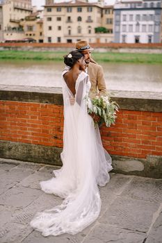 African-American bride and Caucasian groom stand hugging on the embankment of the Arno River, overlooking the city and bridges. Interracial wedding couple. Wedding in Florence, Italy.